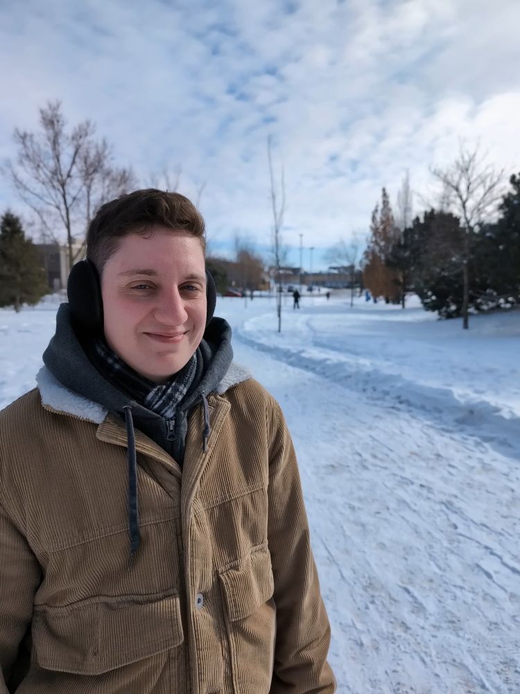 Pic of me, a trans guy, smiling outside in a park full of snow