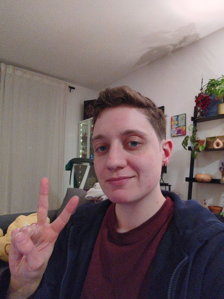 Selfie of me, a trans guy with short hair, with a shy smile, doing a peace sign with my right hand