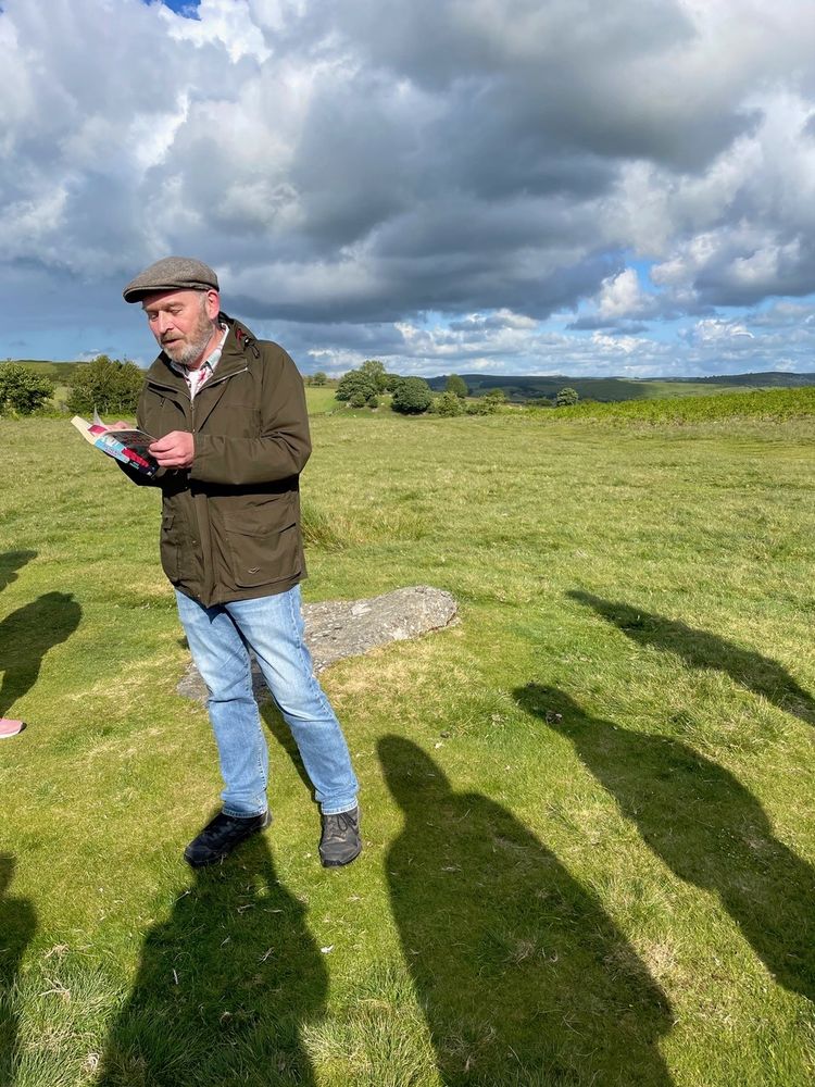 A man reading aloud in a stone circle, with a semi-circle of shadows around him.
