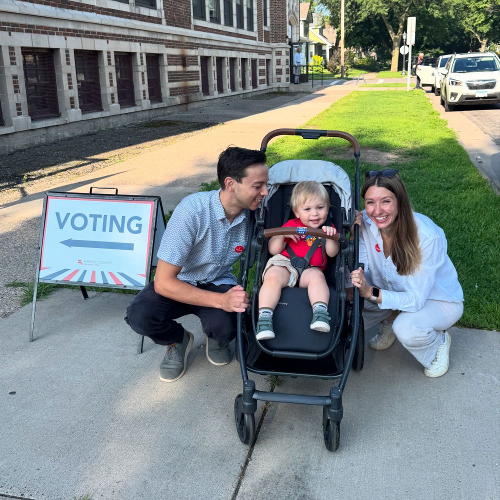 Leo smiles in a stroller while Molly and David crouch next to him, smiling, next to a “voting” sign.