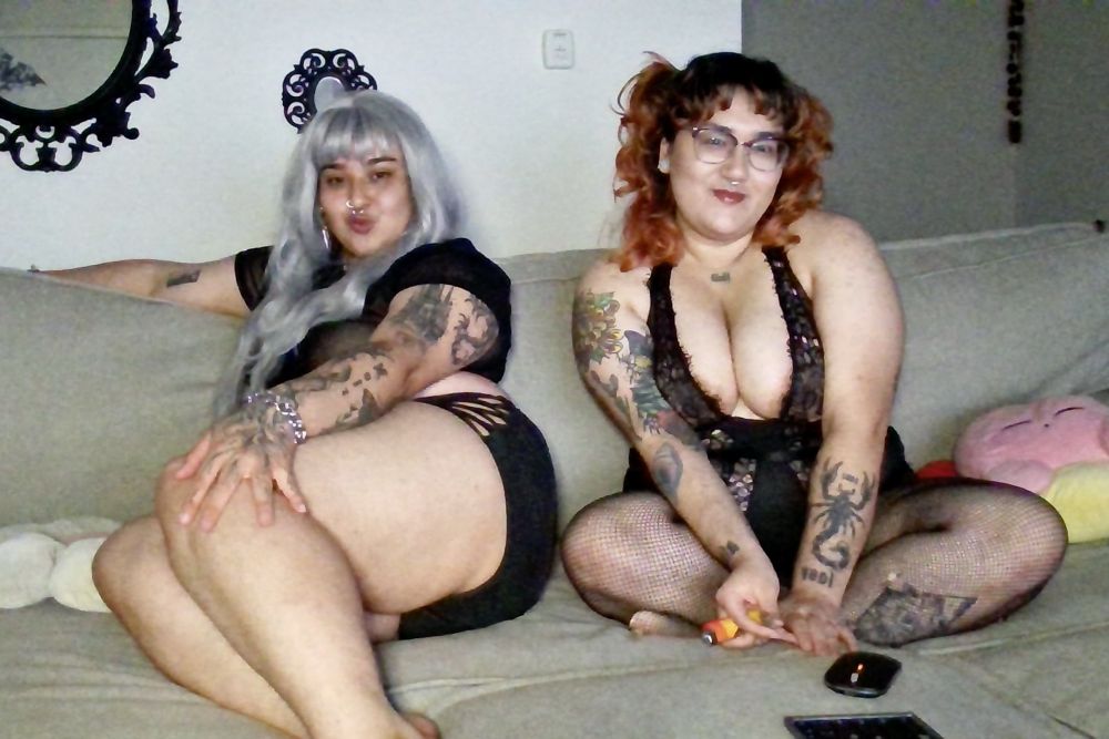 @yosoytudaddy (bambi) on the left wearing black two piece showing legs and kissy face Ele’s Elixir on the right wearing black fishnets and a black bodysuit and a soft smile. they are sitting on a couch  