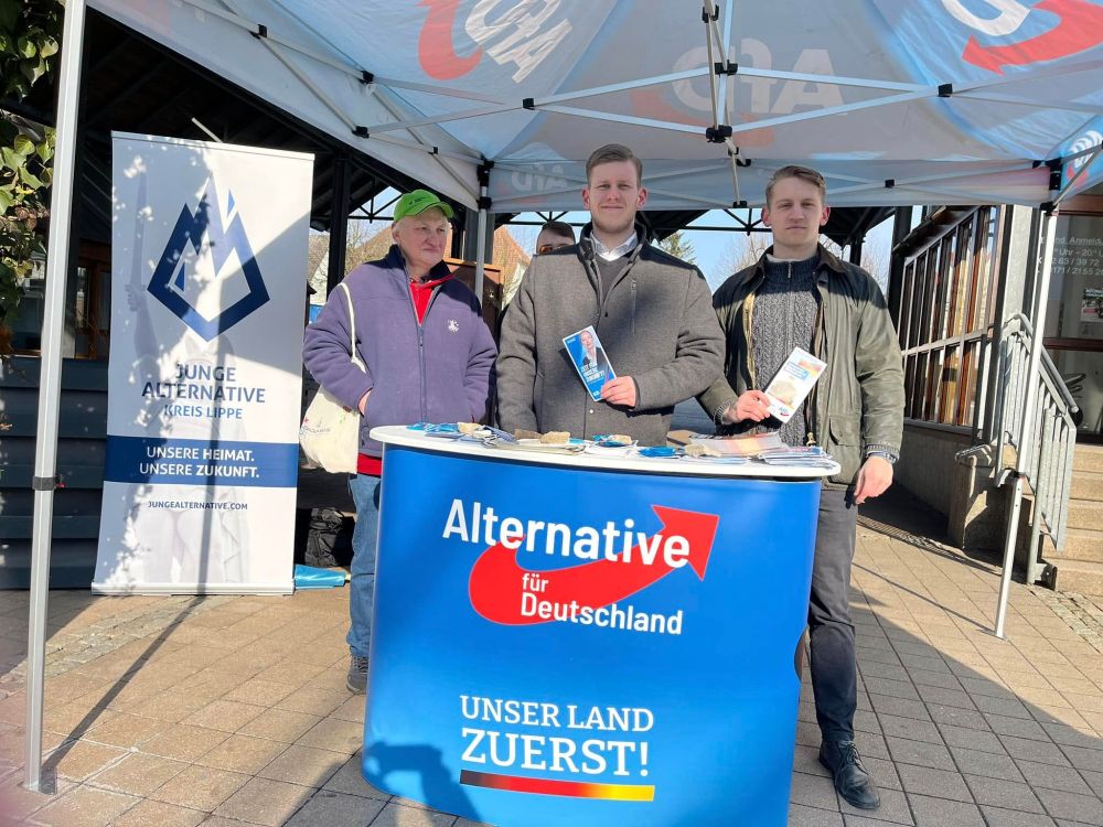 Zu sehen ist ein Straßenwahlkampfstand der AfD Lippe im Februar 2025 in Bad Salzuflen. Im linken Bildhintergrund ist ein Aufsteller der Parteijugendorganisation Junge Alternative zu sehen. In der Bildmitte stehen 3 Männer an einem AfD-Aufstelltisch, in der Mitte Felix Herman Hampe, rechts daneben ein Neonazi der Aktion Hermannsland. Beide halten Flyer der AfD und posieren für das Foto.