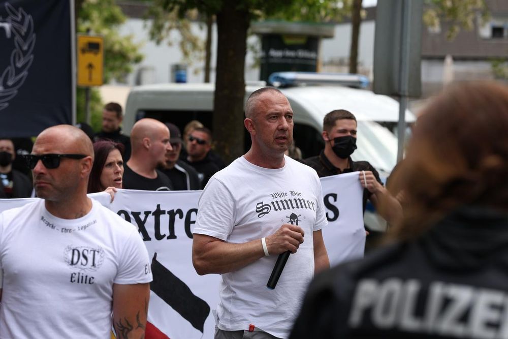 17.05.25 Herford: Bild von der Neonazi-Demo. Im Hintergrund sind Neonazis hinter dem von Daniel Kokott gemalten Fronttransparent mit der Aufschrift "Gegen Linksextremismus - Für Recht und Ordnung" zu sehen. In der Bildmitte ist Daniel Kokott mit geöffnetem Mund zu sehen. In der Hand hält er auf Brusthöhe ein Mikrofon. Links im Bild Vordergrund ist DST-Neonazi Jan zu sehen. (Bild von pixelarchiv)