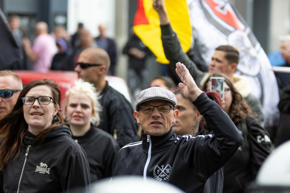 24.05.25: Bild von der kurzen Neonazi-Demo in Bielefeld. Im Bildfokus sind das Pärchen Jana Lange und Thomas Anders aus Bielefeld. Lange trägt eine schwarze Brille und die braunen langen Haare zum Seitenscheitel. Rechts daneben ihr Partner Thomas Anders, ebenfalls mit schwarzer Brille. Er trägt eine grau-karierte Schiebermütze und eine Jacke der Marke Scumbag-Sportswear, die von Neonazi Maurice Quakernack betrieben wird.  (Bild von Nico Kuhn)