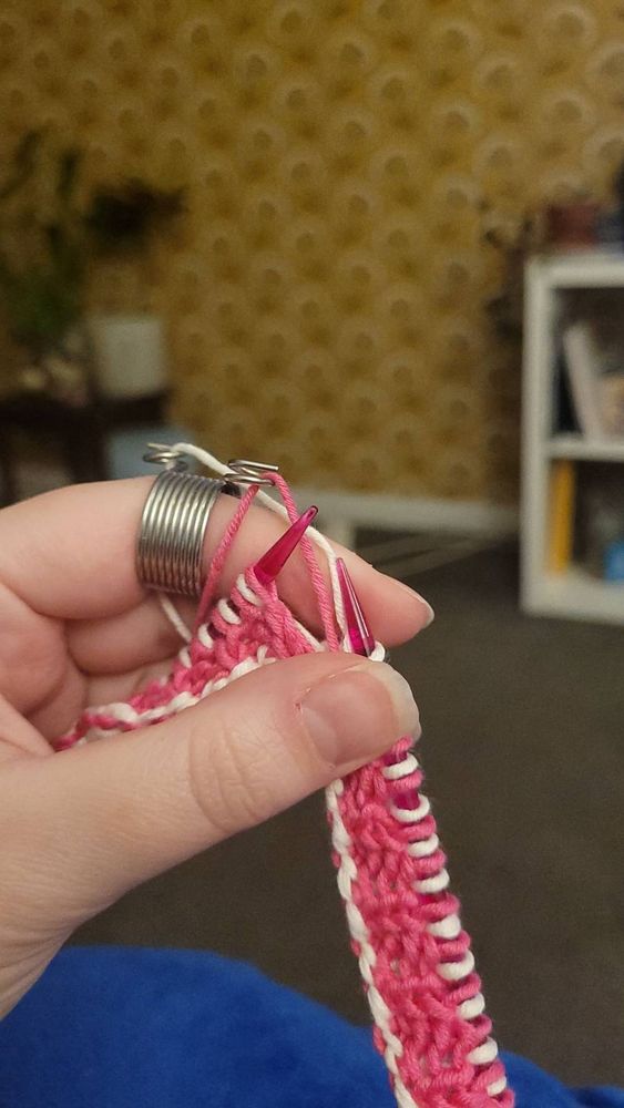 Pink and white knitting project on plastic needles. The strands of yarn are threaded through a wire knitting thimble. 