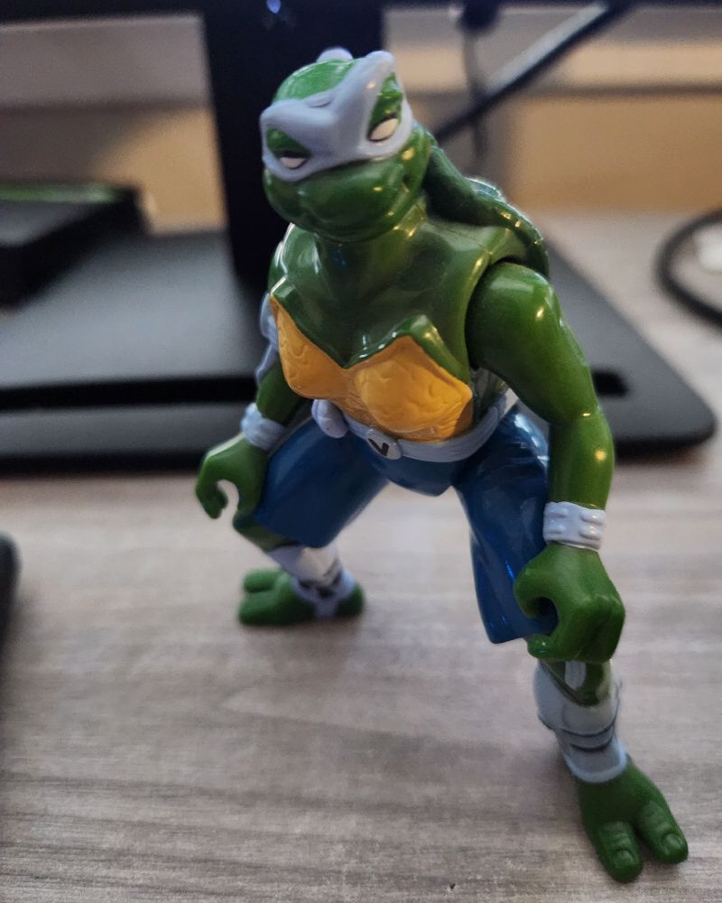 An action figure of Venus de Milo from TMNT: The Next Mutation standing on a beige desk. She's an anthropomorphic turtle mutant with green skin and a yellow chest plate wearing a baby blue mask, belt, armbands, and ankle bands. She's also wearing blue shorts, a belt buckle with a letter V on it, and her mask is styled into a long braid in the back. She has a wide smile and one raised eyebrow, a daring expression.