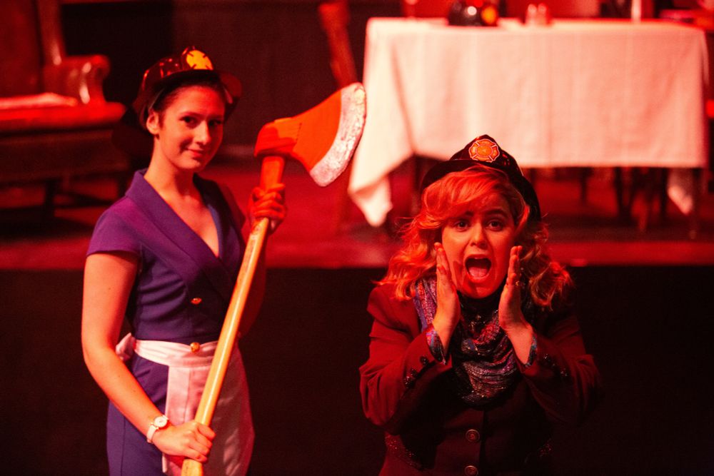 A female firefighter holds an ax ominously and smiles in the background. In the foreground, a woman wearing a fire department helmet screams and looks afraid
