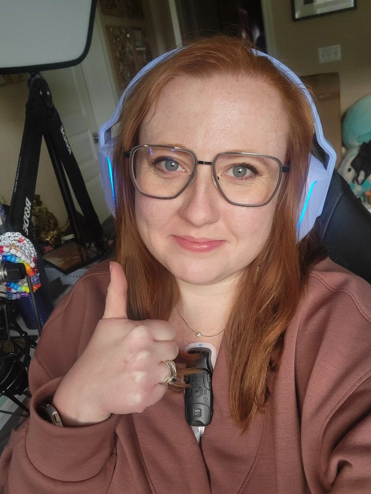 Arcane is shown giving a thumbs up to the camera wearing glasses and purple gaming headphones. She's wearing a clay-colored V-neck shirt that shows a 3 inch tall, 1 inch wide heart monitor on the center of her chest