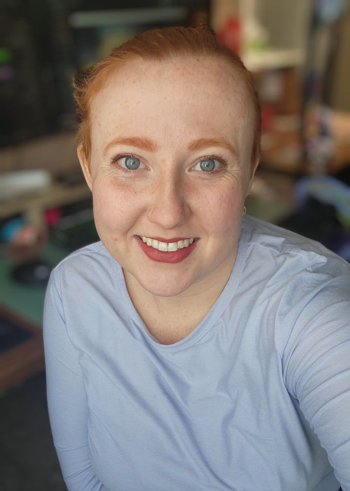 Photo of Arcane, who has blue eyes and red hair, which is up in a ponytail. She is wearing a long-sleeve lavender shirt 