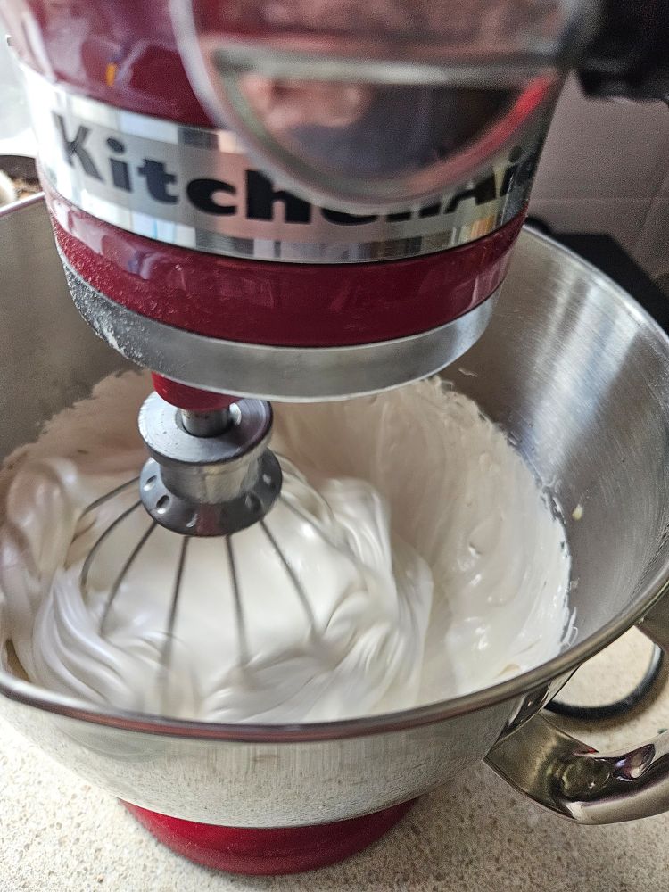 A close up of a red KitchenAid stand mixer with a whisk attachment. The bowl is filled with white cream which is starting to clump around the whisk. Marshmallow! 