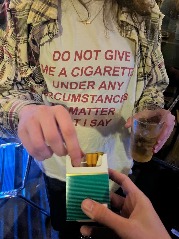 Person wearing a T-shirt that says DO NOT GIVE ME A CIGARETTE UNDER ANY CIRCUMSTANCES NO MATTER WHAT I SAY while holding a beer glass on one hand reaching for a cigarette from a stranger with the other