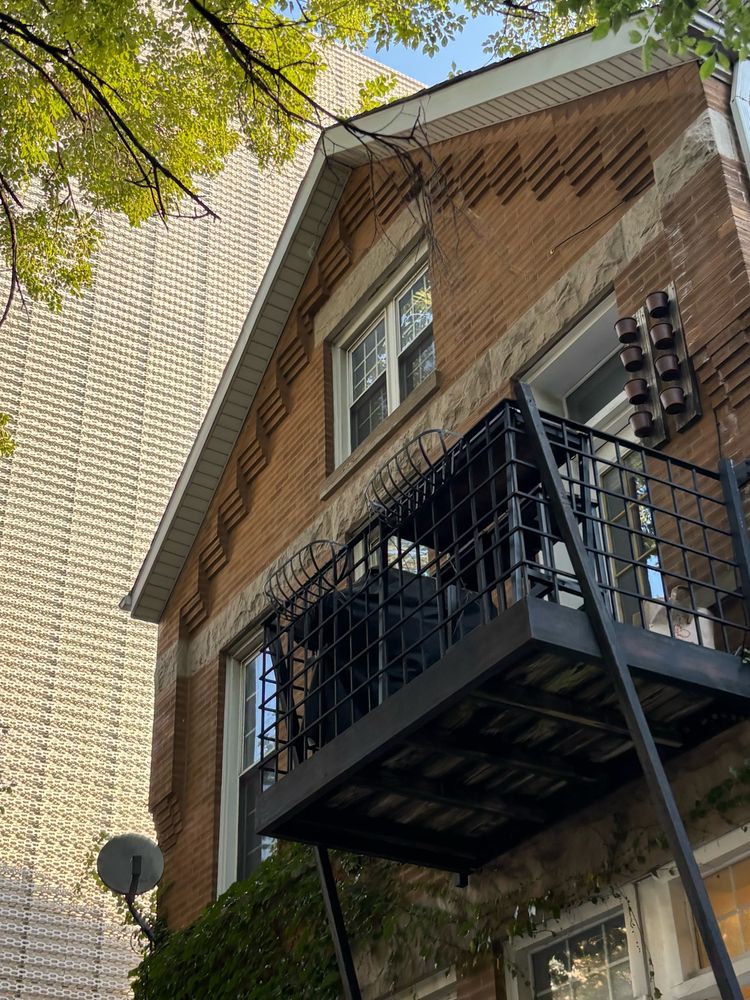 A light-orange-brown brick residence (a workers cottage?) with a raised porch supported by black beams is visible in the foreground, with the strange & somewhat brutalist textured facade of a taller structure in the background. 