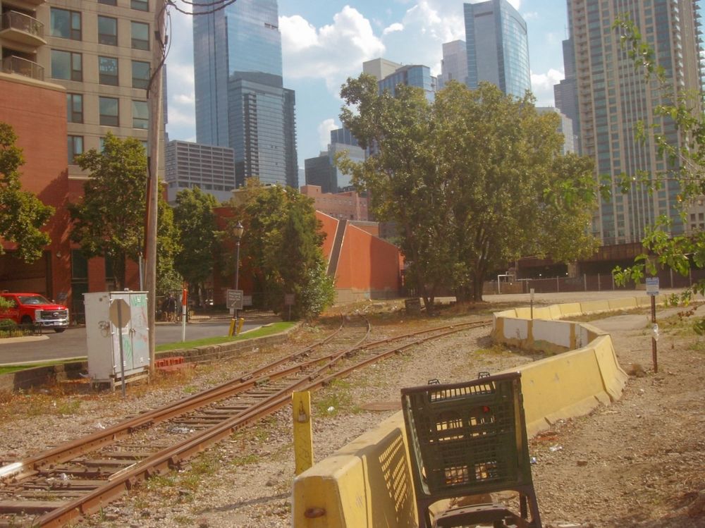 An abandoned shopping cart rests against a barrier beside railroad tracks, which branch off into two directions in the near distance. Buildings are visible in foreground, skyscrapers in background