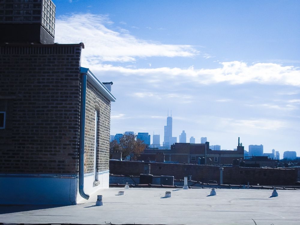 the Chicago skyline is seen in a blue-toned haze from a rooftop in LP area, with several brick structures of varying uses/forms seen in the foreground.  
