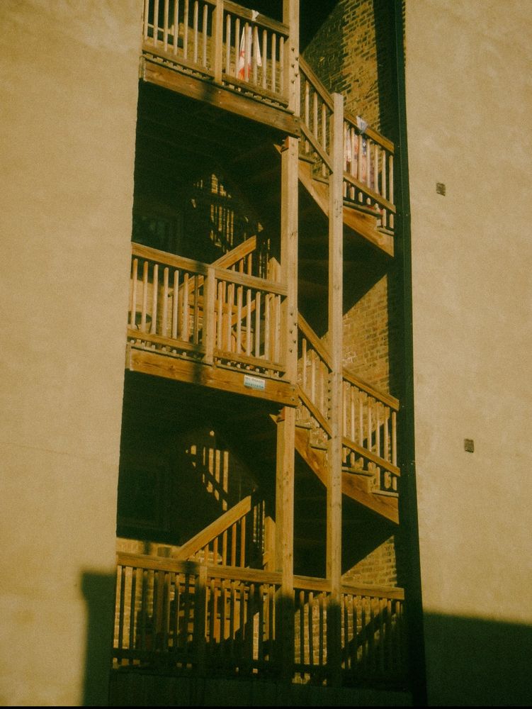 A wooden, external (fire escape?) stairwell zig zags vertically upwards through its exposed nook in between brick and stone walls.  