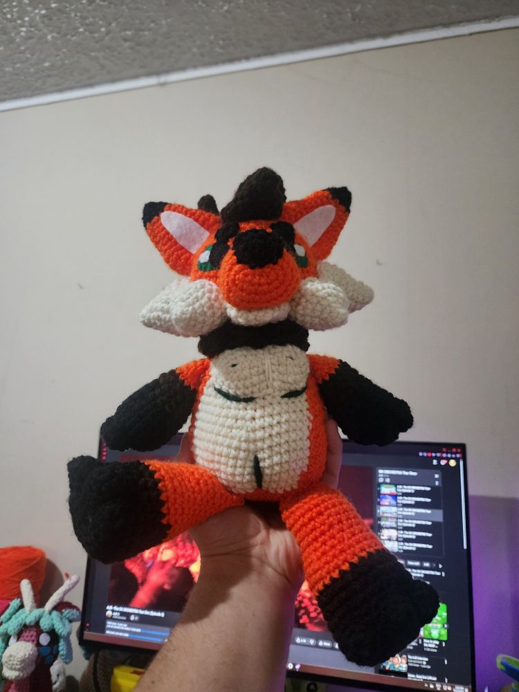 A crocheted plush toy depicting a chubby anthropomorphic fox-bear hybrid creature. It has orange, white, brown, and black fur and green eyes. The plushie has a depiction of two horizontal scars under the chest and one vertical scar on the pelvis, also green.
