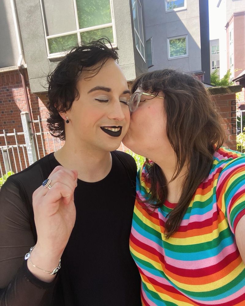 Charlie, dressed all in black with black lipstick has her eyes closed and is smiling, while Sarah, wearing a rainbow striped tshirt kisses Charlie on the cheek.