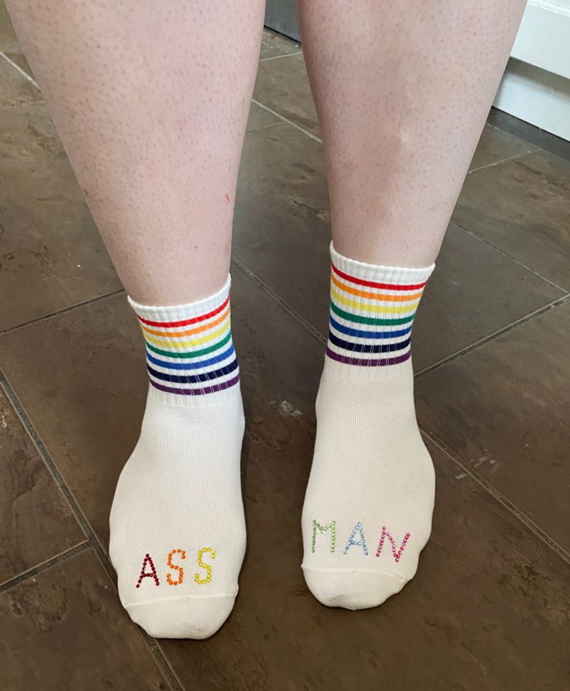 Sarah wearing white socks with rainbow stripes on ankle part of socks, top of socks are bedazzled with “ASSMAN”