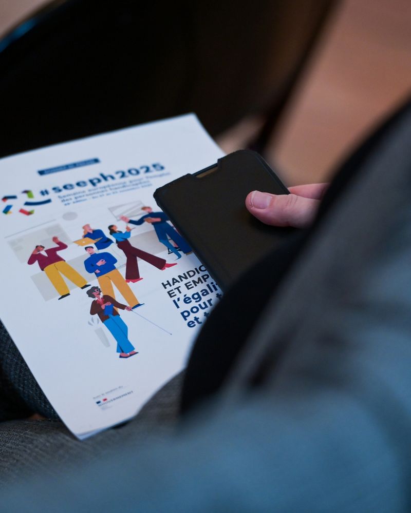 Un plan rapproché montre la main d'une personne tenant un smartphone noir au-dessus d'un document ou d'un programme portant le titre #seeph2025 et illustré par des personnages aux couleurs vives représentant le thème HANDICAP ET EMPLOI : l'égalité pour toutes et tous. 
