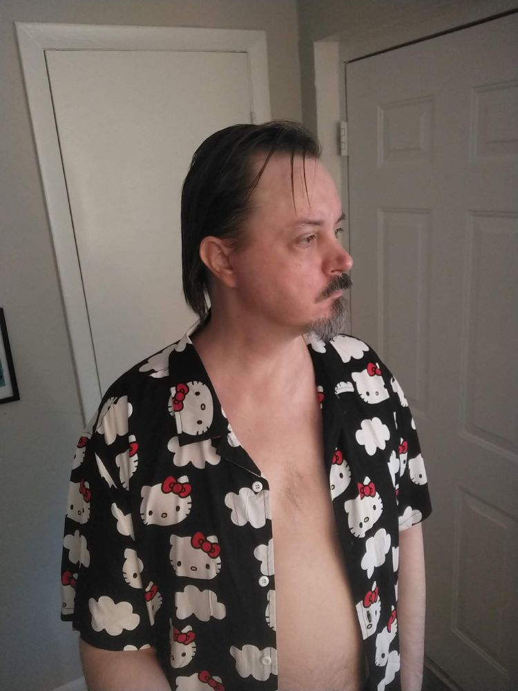 A picture of the poster standing in his apartment. The Hello Kitty shirt from the quoted post hangs from his shoulders, unbuttoned and slightly open.