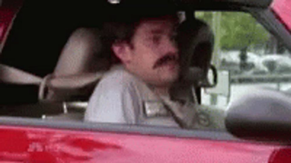 a man with a mustache is sitting in a red car looking out the window .