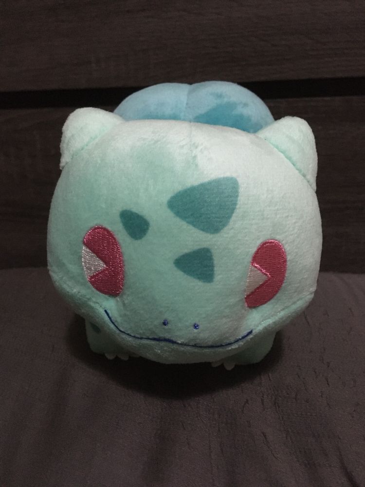 A Bulbasaur Saiko Soda plushie sitting on a grey pillow