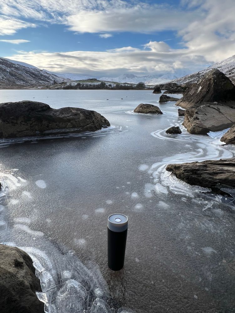A travel coffee cup sat on a frozen lake in front of snow covered mountains and blue skies. 
