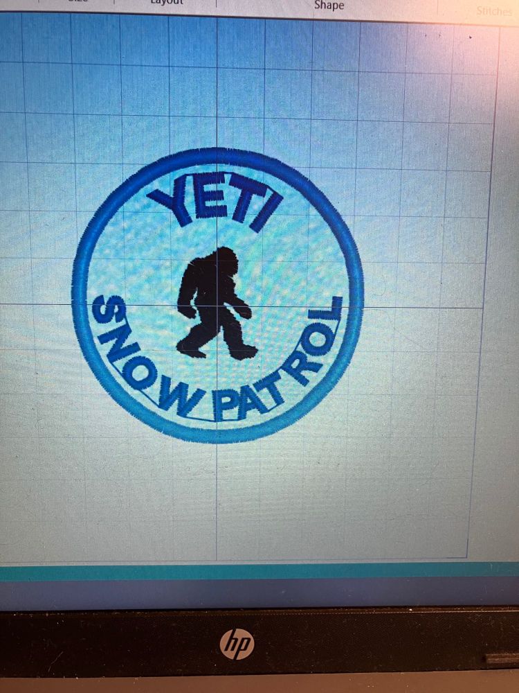 A photo of a laptop screen, showing embroidery software. The design is a cryptid merit patch, with the words Yeti Snow Patrol and a motif of a yeti in the middle. 