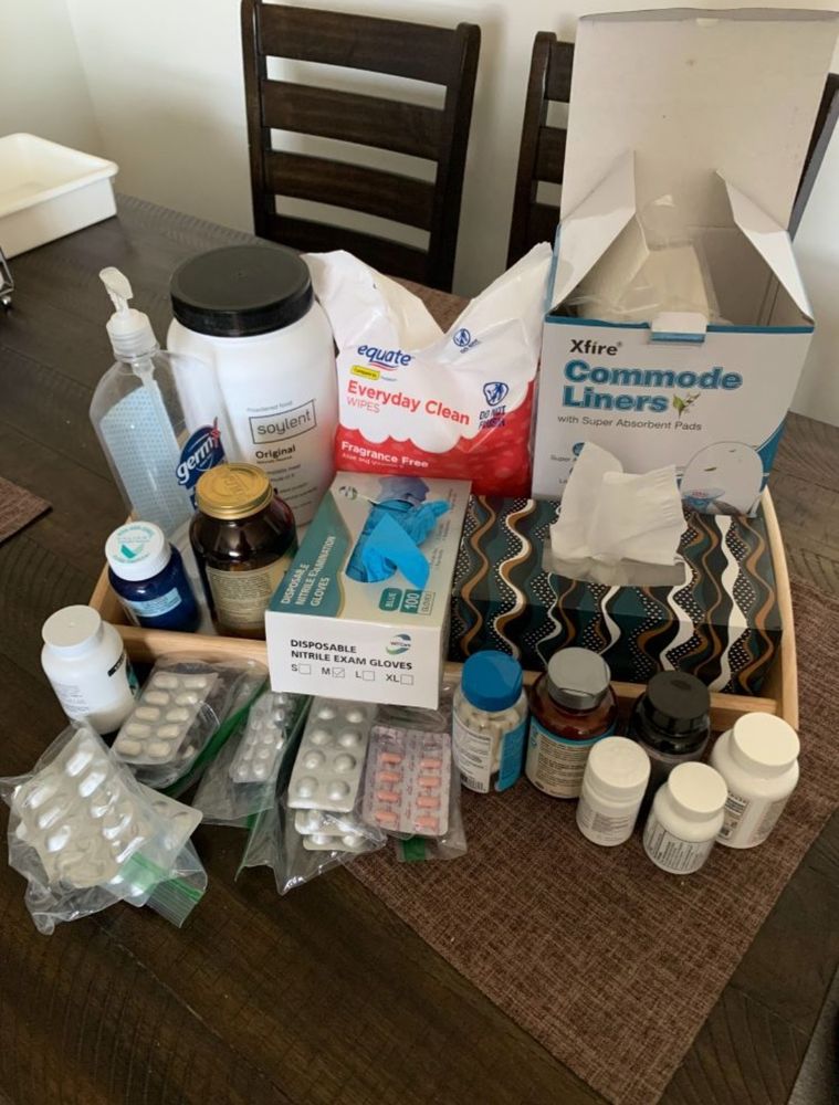 a tabletop medical setup with various health supplies arranged in a neat layout. 

items include multiple bottles of supplements and medications, several blister packs of pills in ziplock bags, a container of Soylent powdered food, a large bottle of Germ-X hand sanitizer, a box of Equate fragrance-free everyday clean wipes, a pack of Xfire commode liners, a tissue box, and a box of disposable nitrile exam gloves. 

the items are placed on a wooden table with two dark wooden chairs in the background. the overall scene suggests preparation for managing chronic illness and caregiving.
