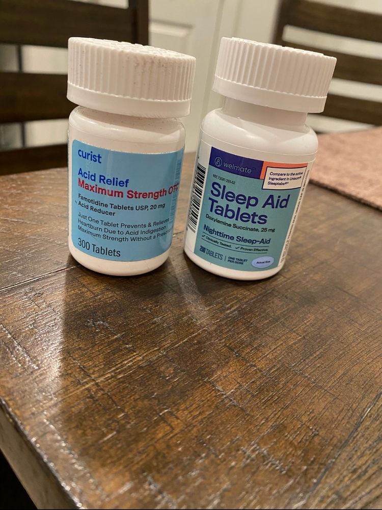 two bottles of over-the-counter medication placed side by side on a wooden surface. 

on the left is a white bottle labeled "curist Acid Relief Maximum Strength OTC," containing 300 Famotidine tablets (20 mg). 

on the right is a white bottle labeled "Wellmate Sleep Aid Tablets," containing 200 Diphenhydramine Succinate tablets (25 mg), a nighttime sleep aid.