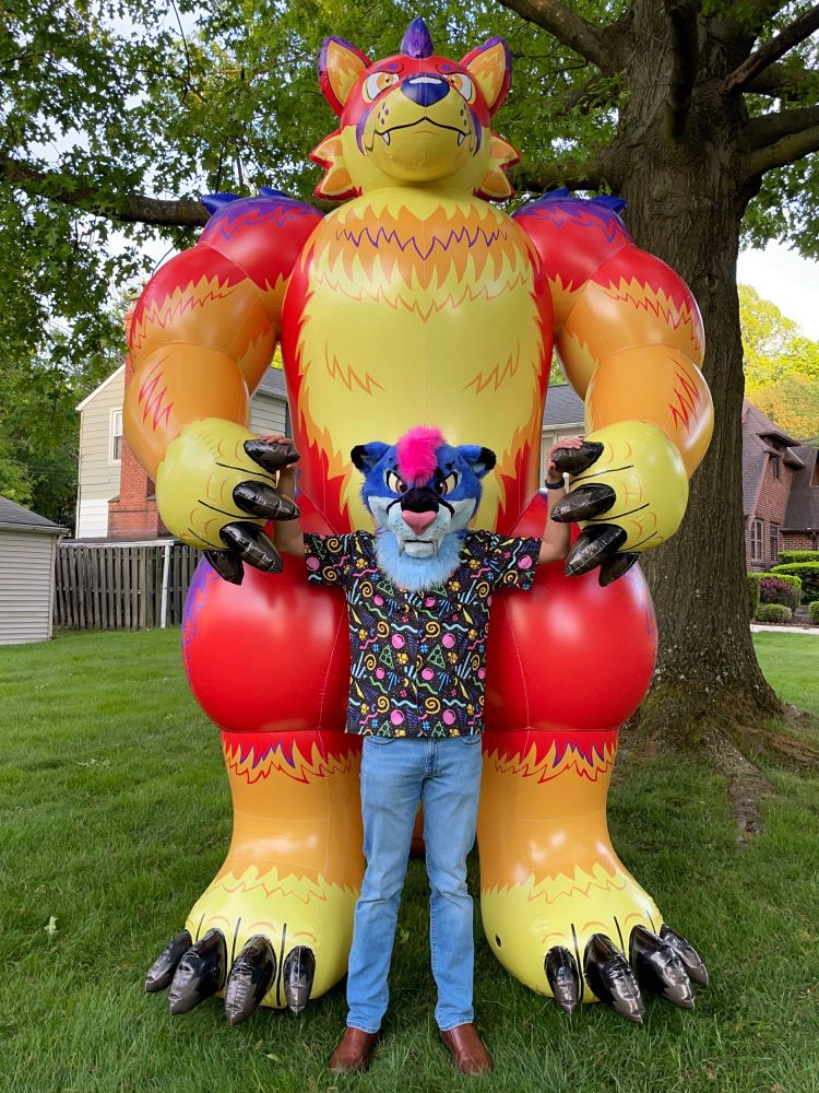 A blue sabertooth in an impossibly busy arcade carpet shirt stands in front of a giant thermal-colored inflatable werewolf under an oak tree.