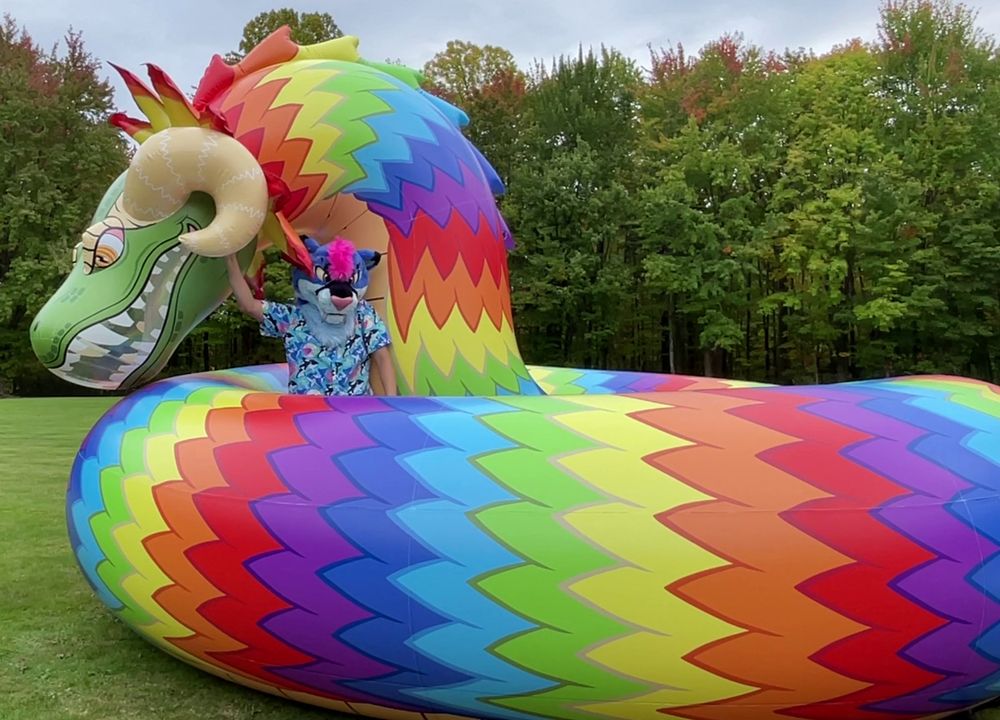A photograph of someone in a blue smilodon partial and loud pooltoy shirt posing with a rainbow-patterned feathered serpent with some very thick coils in a park in early autumn.