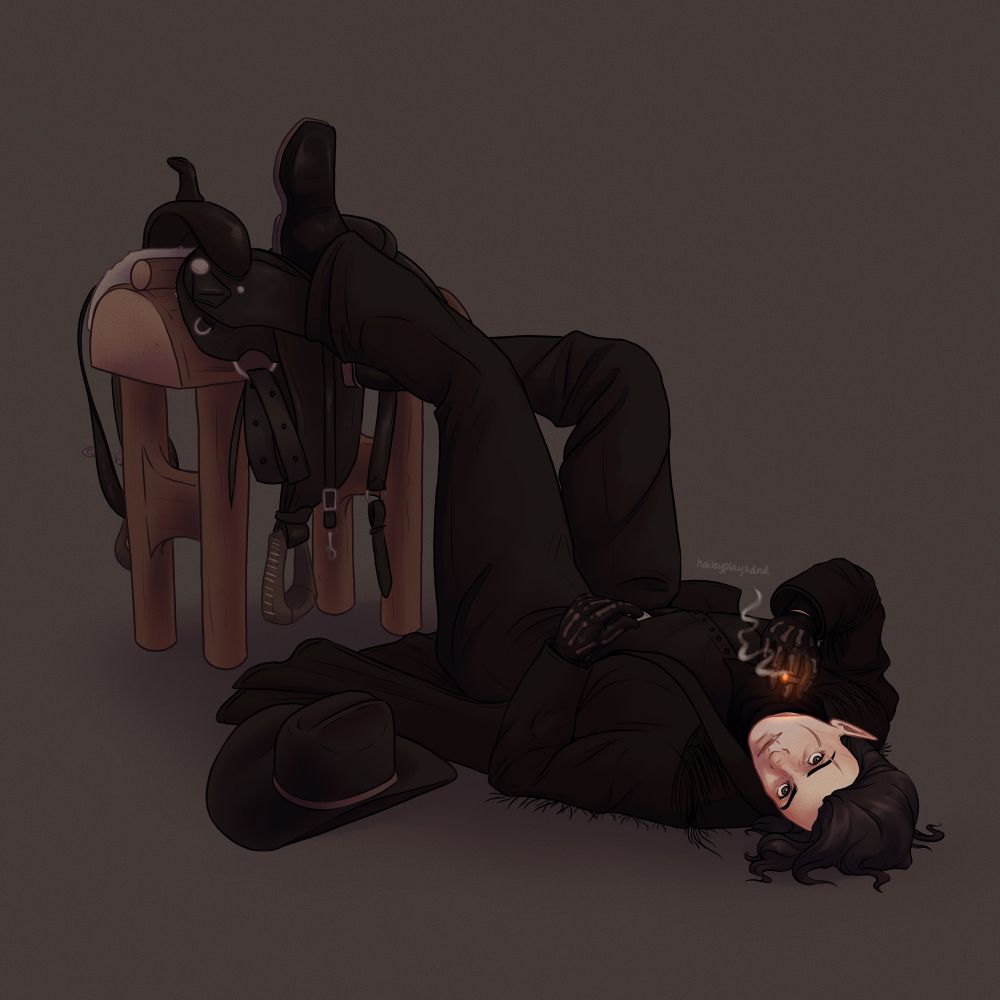 Miss Broody Dark Cowgirl is lounging on the ground, her head tilted back and up toward us, a lit cigarette between her gloved fingers. Her legs are propped up on a black saddle sitting atop a saddle stand. There is a cowgirl hat resting on the ground next to her. The background is a plain muted brown/grey.