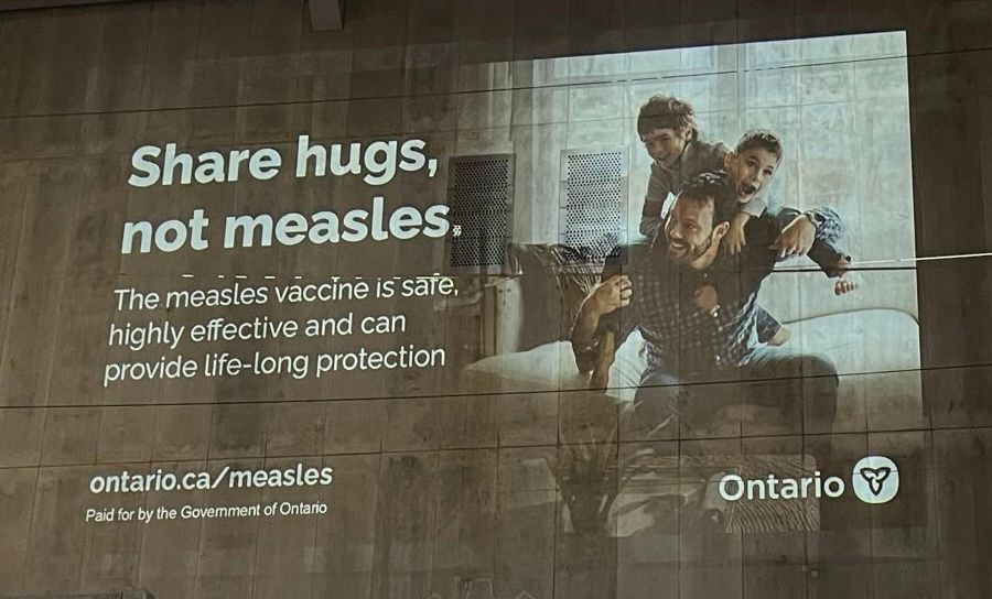 A photo of an advertisement paid for by the Government of Ontario is depicted. The words ‘Share hugs, not measles.’ are in large, bold text on the left side of the ad, followed by the message ‘The measles vaccine is safe and effective and can provide life-long protection.’ On the right side of the add is an accompanying image of a man seated on a couch, with 2 young children climbing on his back. Everyone appears to be having fun, and no one has an obvious rash. 

At the bottom, a url is provided to the site Ontario.ca/measles, and the Ontario government logo is displayed. 