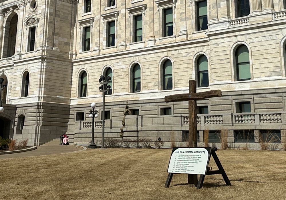 Closer up photo taken March 25 that shows the Minnesota State Capitol’s facade with a display in the foreground of a wooden cross planted in the ground with a sandwich-board sign with the Ten Commandments printed on it. 