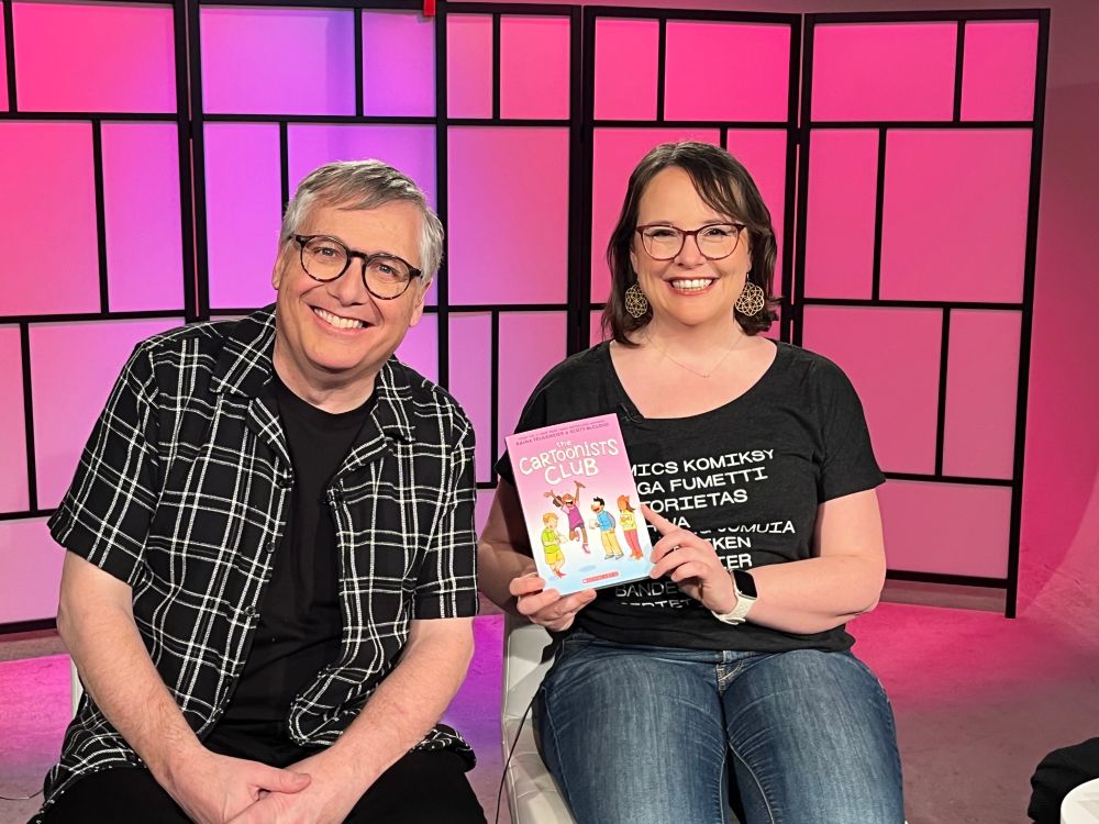 Scott McCloud and Raina Telgemeier showing off THE CARTOONISTS CLUB. They’re both dressed in black and white, in front of a pink and black background evoking comic book panels. 