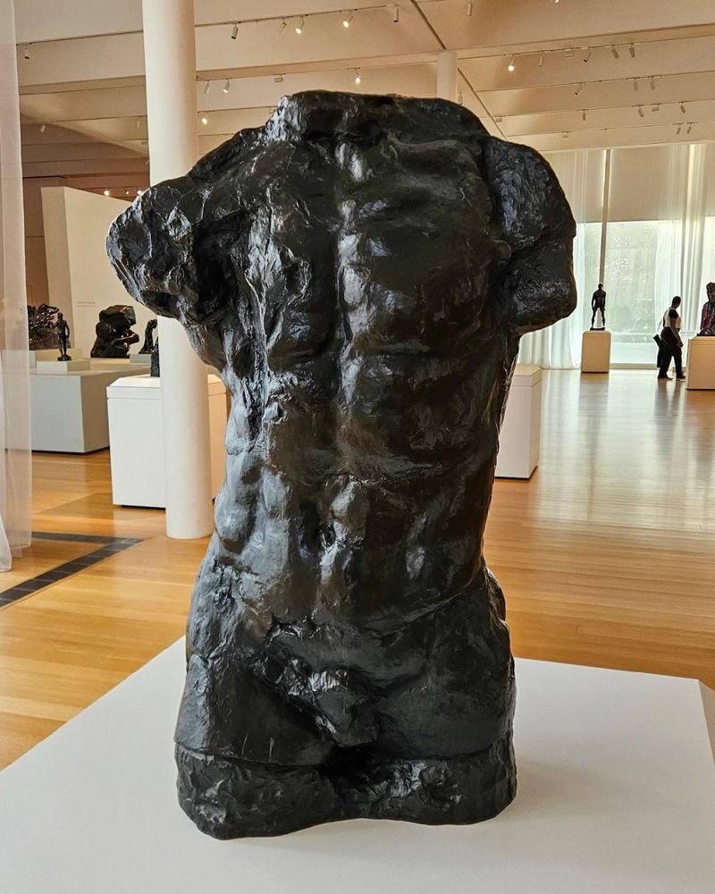 Bronze torso sculpture displayed in a museum gallery with other artworks and visitors visible in the background.