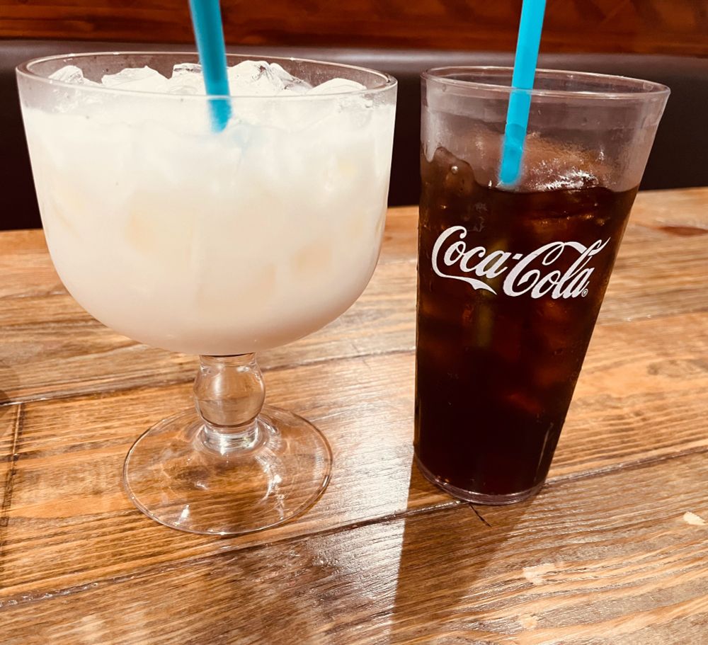 A very large and wide glass of horchata. It is next to a regular glass of soda for a size comparison. The horchata is much bigger than the soda!