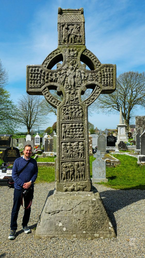 A picture of me stood next to the early medieval stone high cross known as Muiredach’s Cross at Monasterboice on a bright sunny day