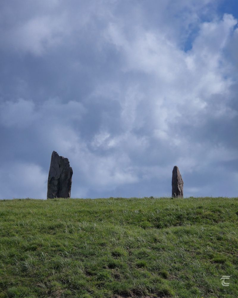 A pair of standing stones atop a green hill under a cloudy sky