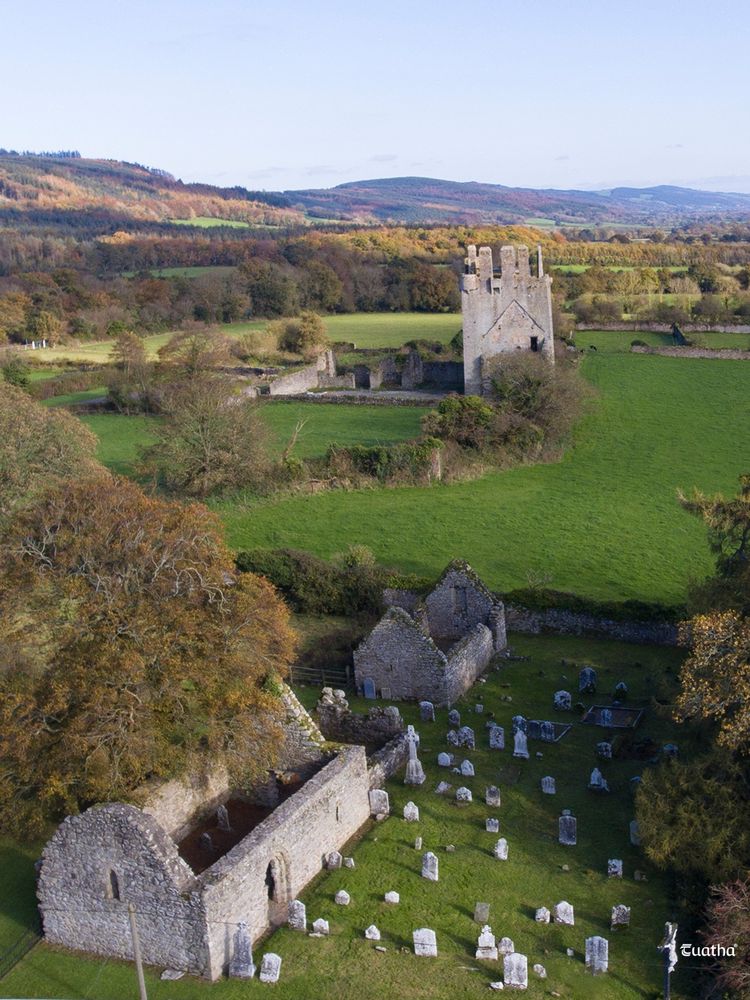An aerial view of the small 12th century romanesque church surrounded by a graveyard, with Kilcash Castle in the background.