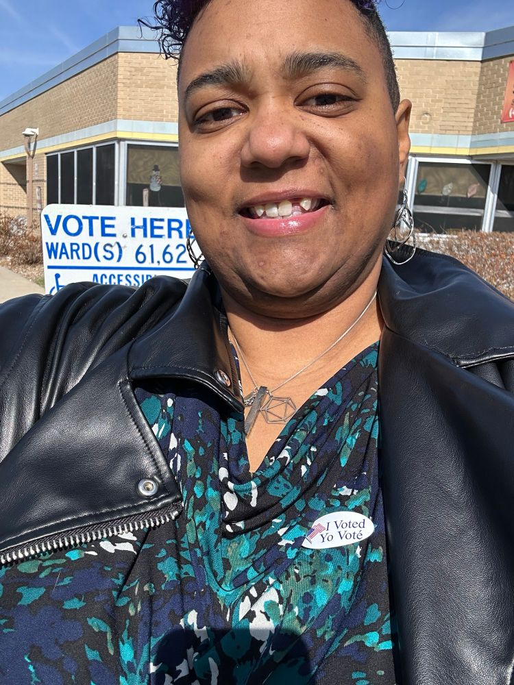 Jessiye wearing a green and blue blouse with a sticker that says I voted in a black leather jacket and it’s very sunny outside as she stand outside a voting location
