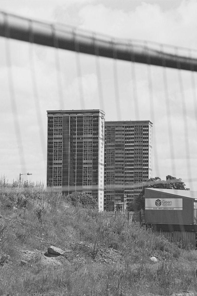 A black and white photograph of two high rise buildings behind a metal wire fence with some waste land in the foreground. 