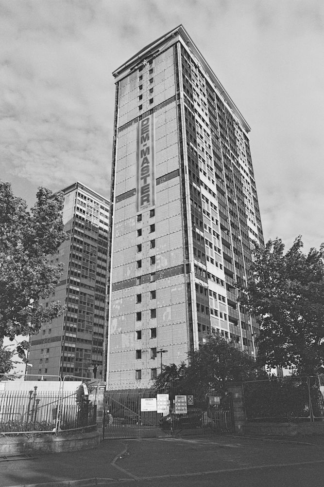 A black and white photograph taken on 35mm film of two high rise buildings that have been prepped for demolition. The most prominent building in the photo has a Dem-Master banner running down the side of it, which is the company carrying out the demolition. The buildings are behind a metal gate and white fence. These are at the bottom of the frame with trees either side of the frame. 