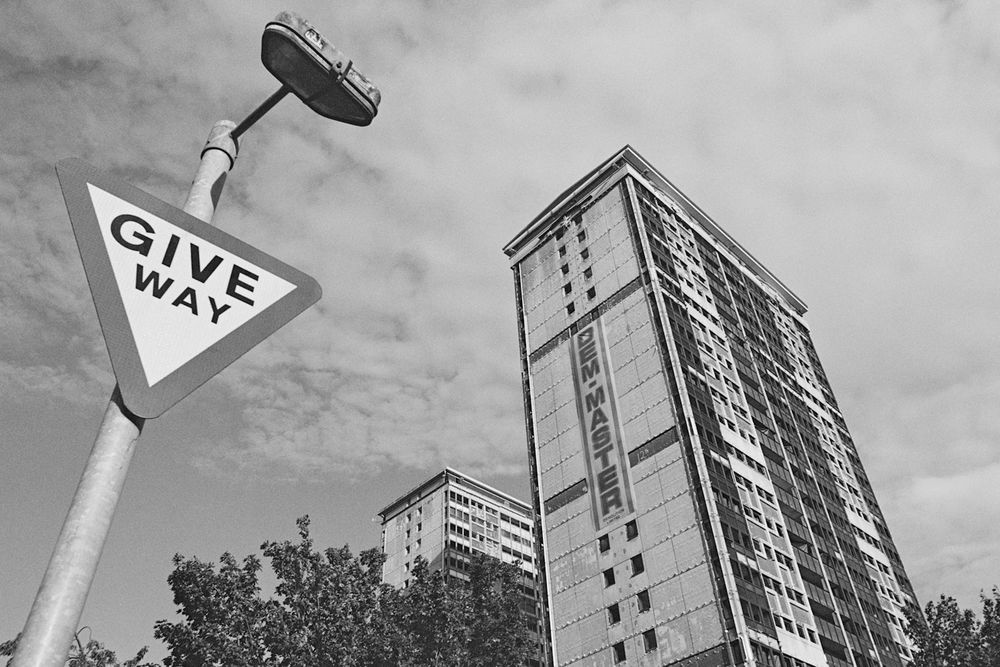 A black and white photograph taken on 35mm film looking up at two high rise buildings on the right of frame and a "give way" road sign to the left of the frame. The high rise buildings are prepped for demolition with a banner running down the side of the most prominent and closest building in the image. The banner reads "Dem-Master" which is the company carrying out the demolition work to these two high rise buildings. 