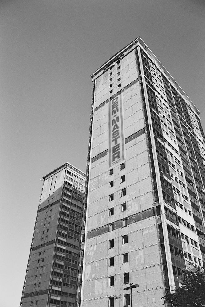 A black and white photograph taken on 35mm film, of two high rise buildings stretching up into the sky. These buildings have been prepped for demolition and there is a large banner down the side of the nearest building which has "DEM-MASTER" written on it. This is the name of the demolition company carrying out the work. 