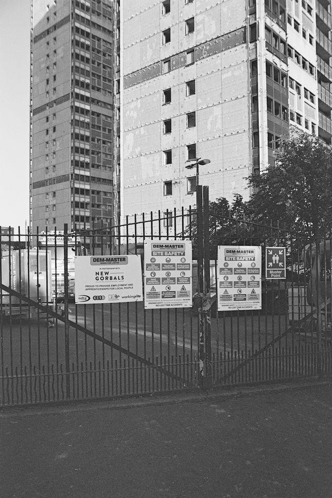 A black and white photograph taken on 35mm film, of a black metal fence with warning/safety signs on it giving information about the hazards present behind the gate. There are sections of two high rises in the frame behind the metal gate. These buildings have been prepped for demolition. 