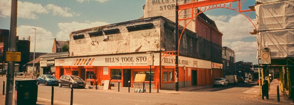 A panoramic photograph taken on colour 35mm film of Bill's Tools, a tool shop/hardware store in Glasgow's famous Barras. The shop is on a corner and with the panoramic view you can see both fronts. The sun is shining down on the scene. The bright red colour of the store pops out along with the metal arch entrance to the Barras which is to the right of the shop. The sky above is blue with white clouds. 