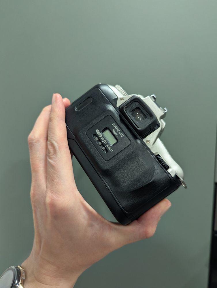 A colour photo of a Nikon F55 35mm SLR camera being held up against a green background. The camera is silver with a black data back. 