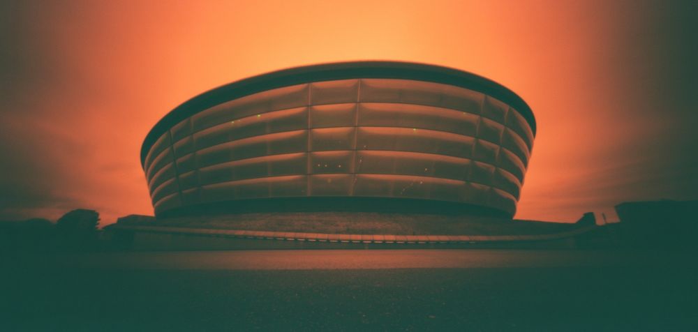 A Redscale 120 film photograph (6x12) of the OVO Hydro venue in Glasgow. The distinctive building looks almost like a crashed flying saucer. The Redscale film means the colours within this photo are various shades of red, orange and yellow. As this was taken with a Holga 120WPC pinhole camera there is a heavy vignette on this image with the Hydro building appearing in the centre of the frame. 